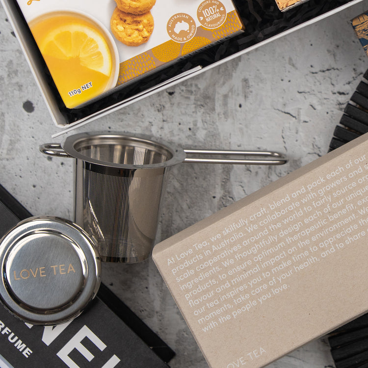 A flat lay of The Hamper Boutique Co's Exotic Explorations Tea Hamper, featuring a tea strainer, a LOVE TEA tin, lemon cookies, and a box with printed text about Love Tea on a gray textured surface.