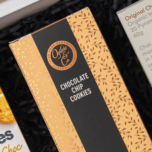 A rectangular box of The Hamper Boutique Co Exotic Explorations Tea Hamper with a gold and black, leaf-patterned design, displayed on a bed of black crinkle paper.