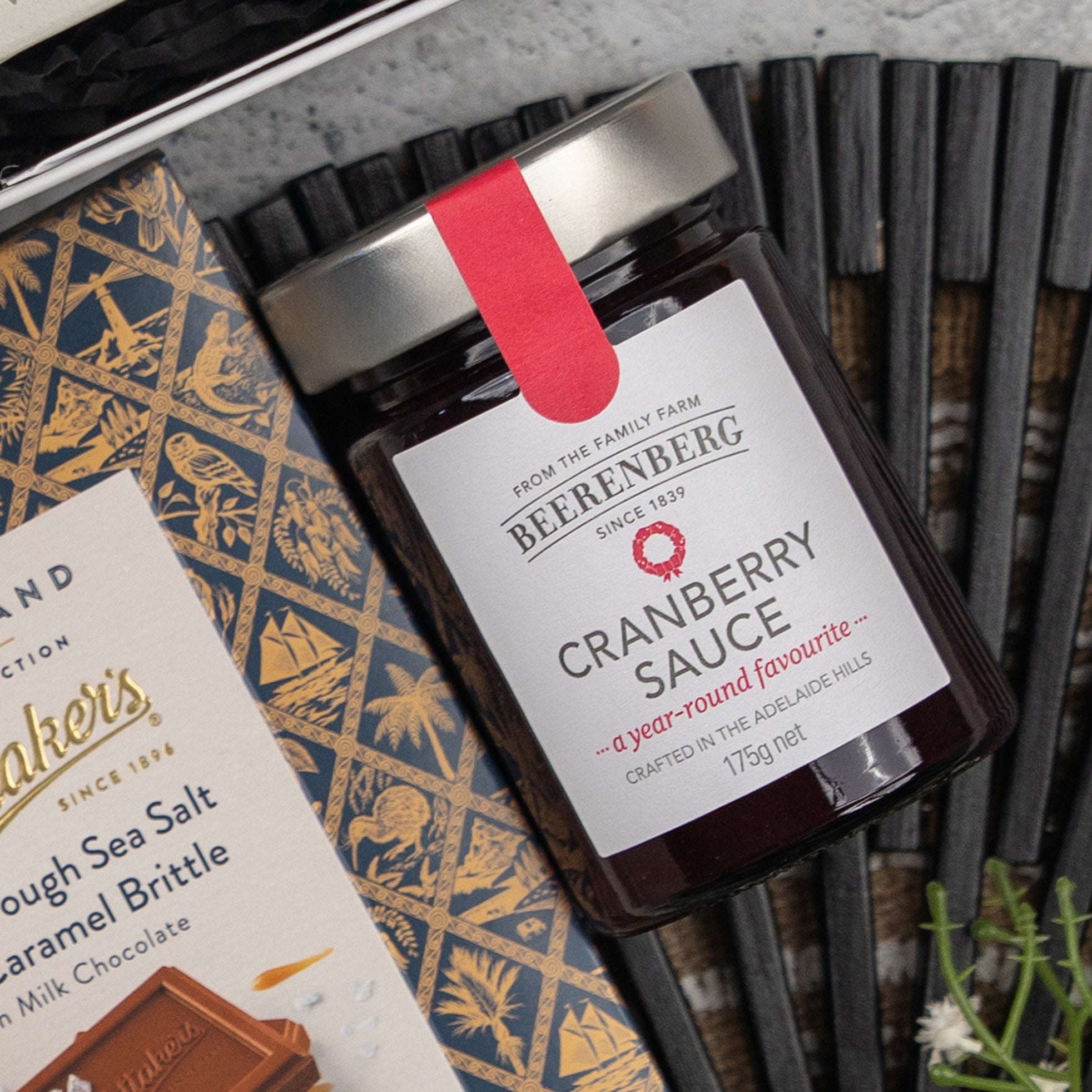 An Exotic Explorations Tea Hamper by The Hamper Boutique Co is displayed on a dark patterned surface, with some white flowers in the corner.