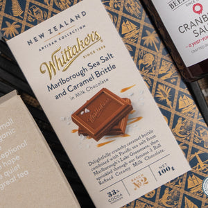 An Exotic Explorations Tea Hamper by The Hamper Boutique Co, in decorative packaging with product details, is displayed on a patterned surface alongside other food items.
