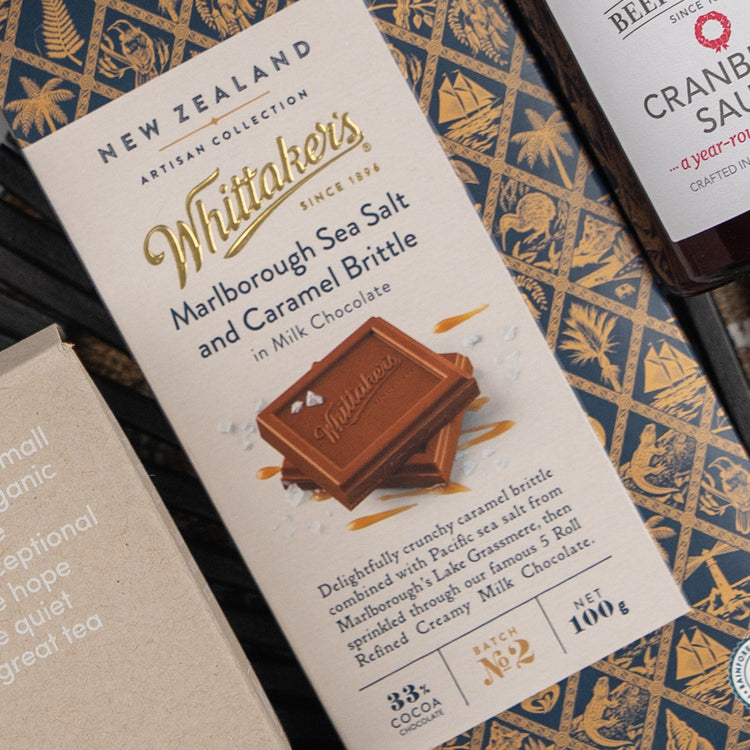 An Exotic Explorations Tea Hamper by The Hamper Boutique Co, in decorative packaging with product details, is displayed on a patterned surface alongside other food items.