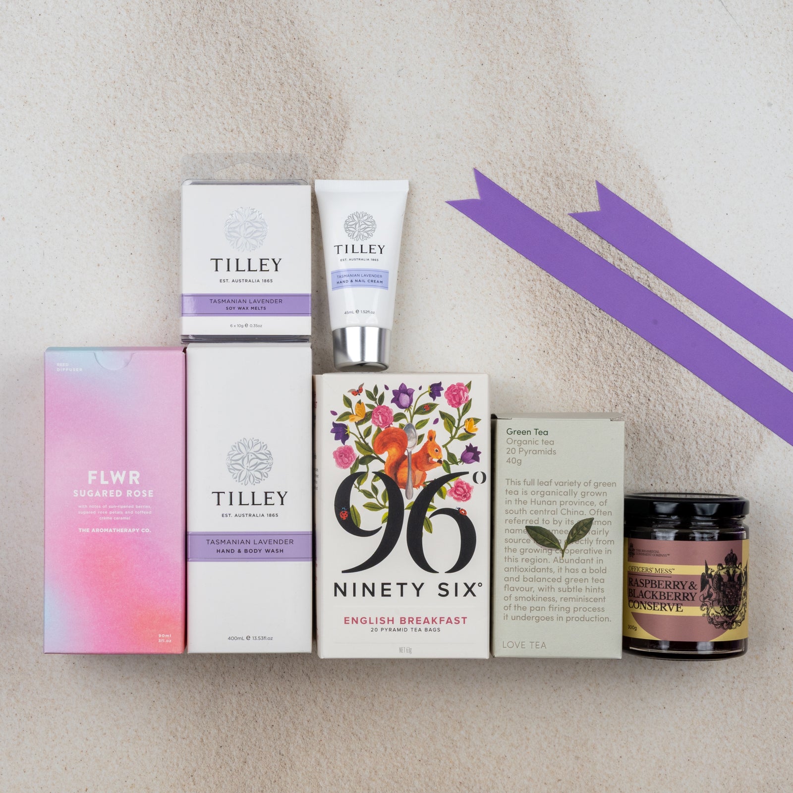 A flat lay of The Hamper Boutique Co's Relax and Rejuvenate Tea Hamper, featuring hand and body wash, cream, perfume, teas, and preserve, arranged on a light surface with purple ribbons.