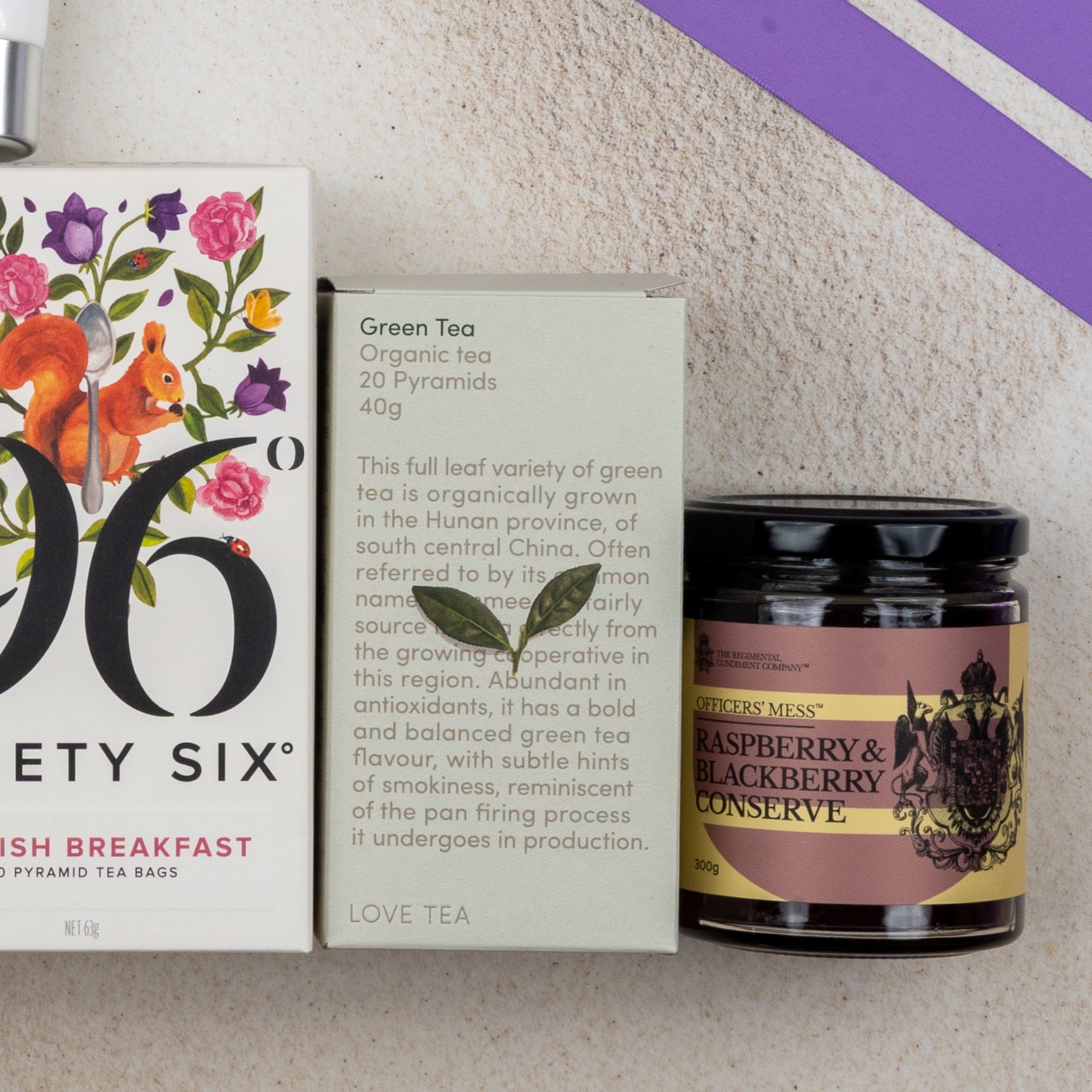 The Hamper Boutique Co's Relax and Rejuvenate Tea Hamper, featuring beautifully illustrated tea boxes and a jar of raspberry & blackberry conserve, is displayed on a light surface with purple diagonal lines in the corner.