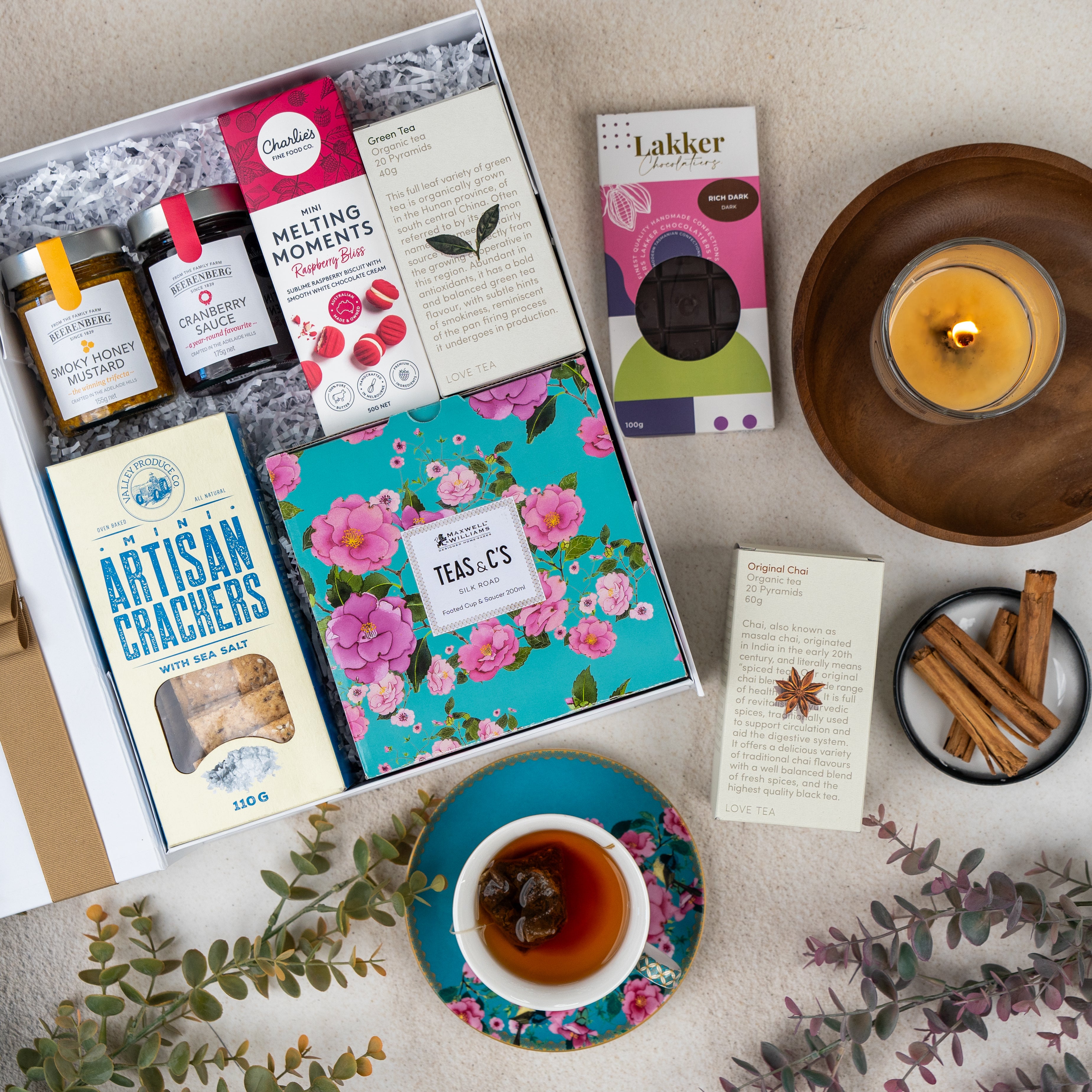 A flat lay of The Hamper Boutique Co's Sweet Indulgence Tea Hamper featuring tea, honey, cookies, crackers, a floral tea tin, a cup of tea on a saucer, cinnamon sticks, a burning candle, and greenery on a beige surface.