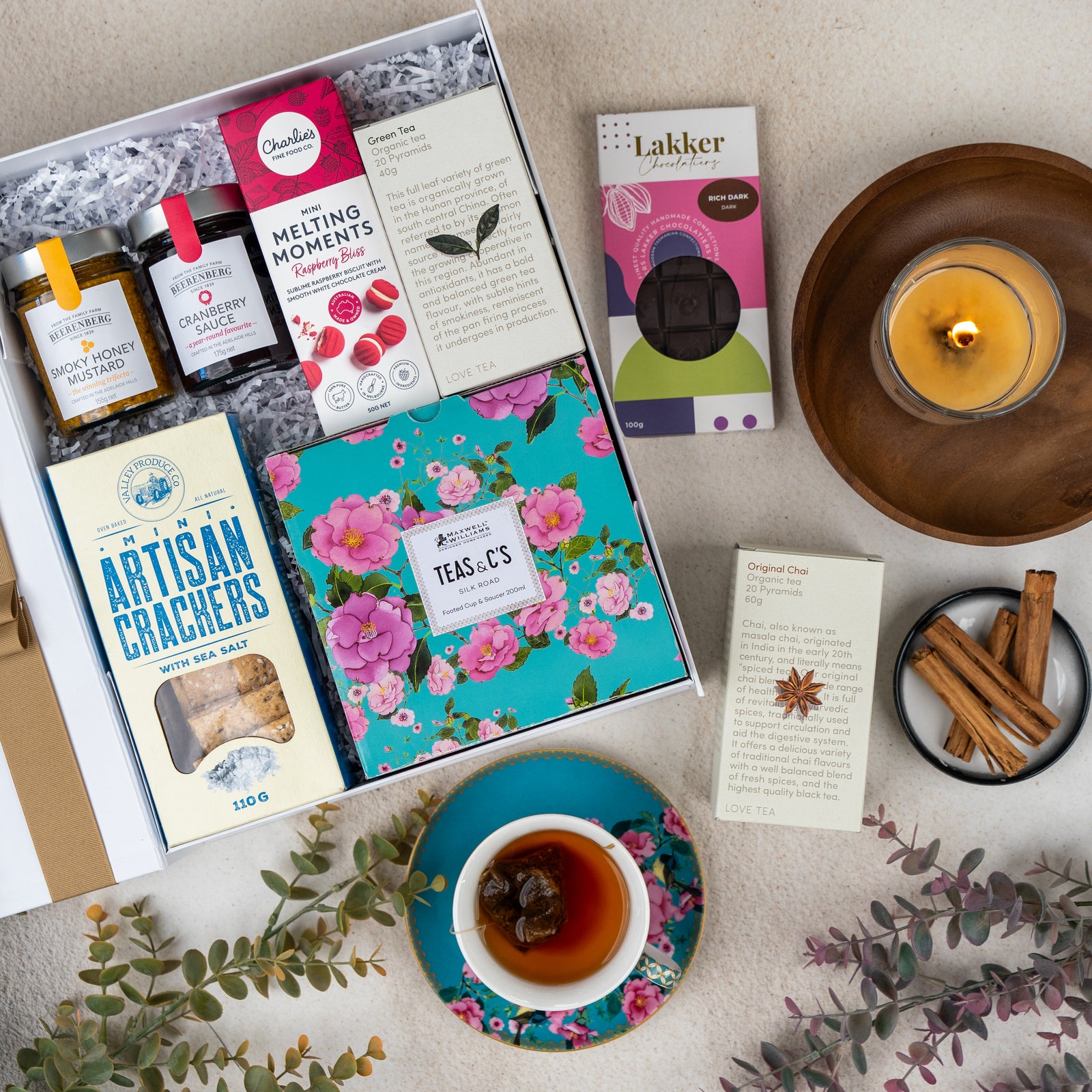 A flat lay of The Hamper Boutique Co's Sweet Indulgence Tea Hamper featuring tea, honey, cookies, crackers, a floral tea tin, a cup of tea on a saucer, cinnamon sticks, a burning candle, and greenery on a beige surface.