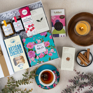 A flat lay of The Hamper Boutique Co's Sweet Indulgence Tea Hamper featuring tea, honey, cookies, crackers, a floral tea tin, a cup of tea on a saucer, cinnamon sticks, a burning candle, and greenery on a beige surface.