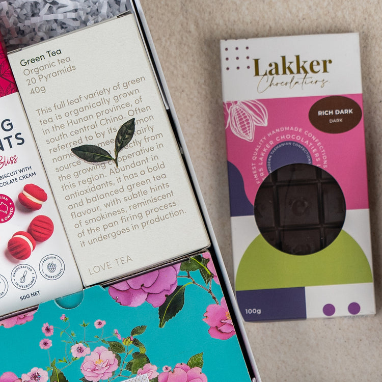 The Hamper Boutique Co’s Sweet Indulgence Tea Hamper, in a floral decorative box, features green tea pyramids and a Lakker Chocolates dark chocolate bar, displayed on a textured beige surface.