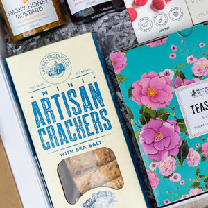 The Hamper Boutique Co’s Sweet Indulgence Tea Hamper includes mini sea salt crackers in blue packaging, a floral teal tea box, shredded paper filling, jars of honey mustard, and other gourmet treats—perfect for gifting.