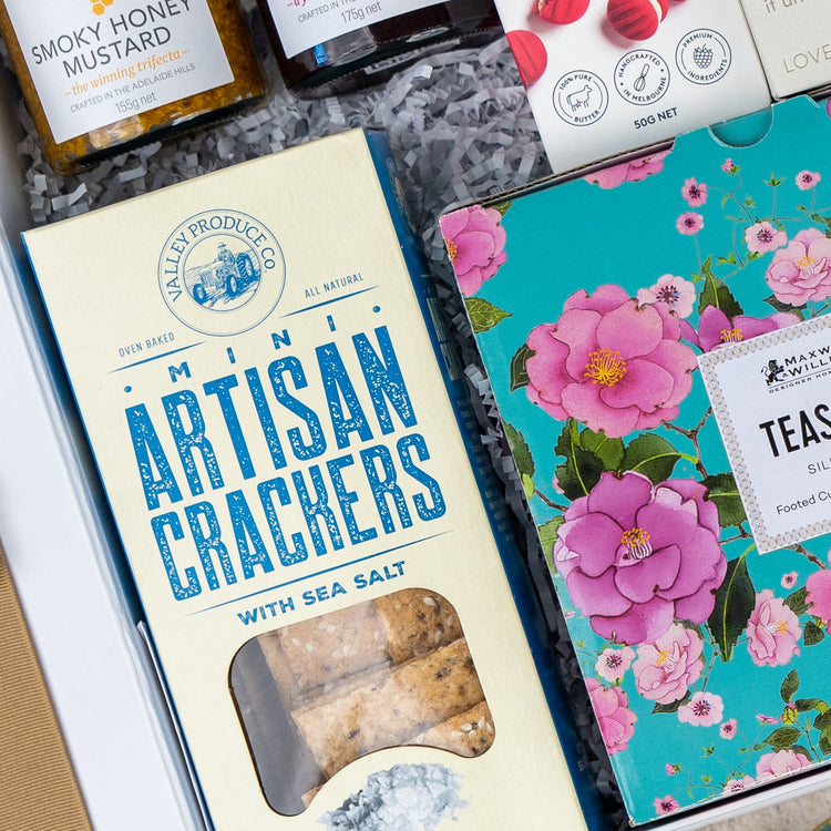 The Hamper Boutique Co’s Sweet Indulgence Tea Hamper includes mini sea salt crackers in blue packaging, a floral teal tea box, shredded paper filling, jars of honey mustard, and other gourmet treats—perfect for gifting.