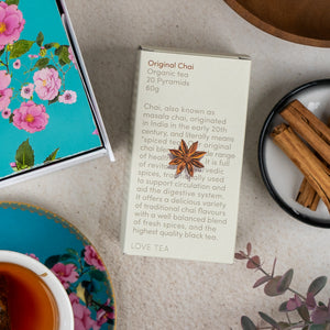 A box of Sweet Indulgence Tea Hamper by The Hamper Boutique Co sits on a table with a star anise on top, surrounded by a cup of tea, cinnamon sticks, a floral tin, and a blue floral-patterned dish.