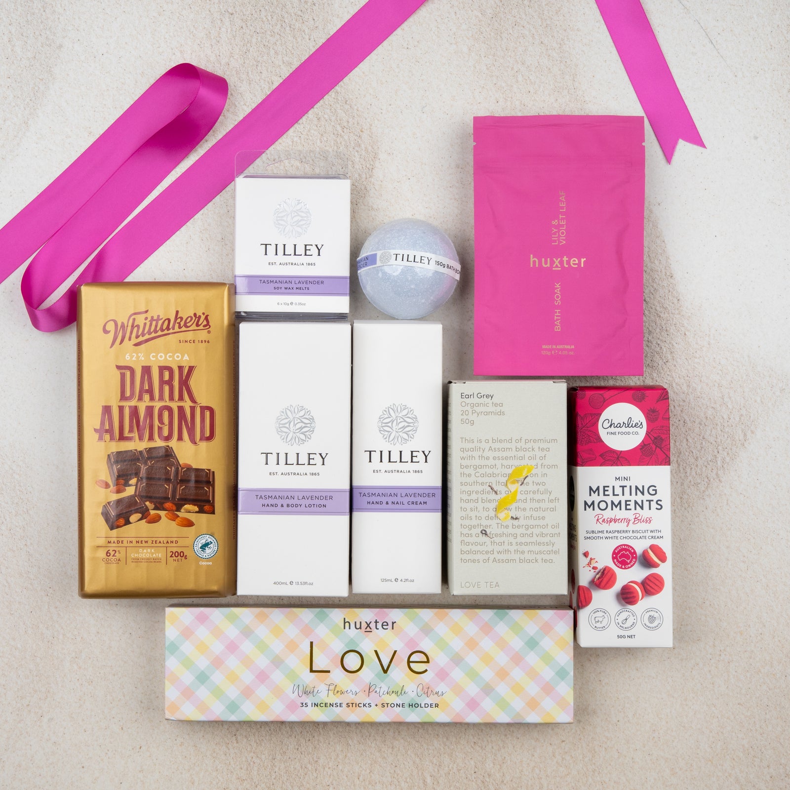 The Hamper Boutique Co’s Rejuvenation and Wellness Hamper sits on a white surface, featuring chocolate, bath products, a pink ribbon, lavender bath bomb, Tilley soaps, Huxter Love incense, and raspberry melting moments cookies.
