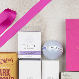 A flat lay of The Hamper Boutique Co’s Rejuvenation and Wellness Hamper on a light surface, showcasing soy wax melts, a bath bomb, a pink ribbon, chocolate bar, and a pink tea package.