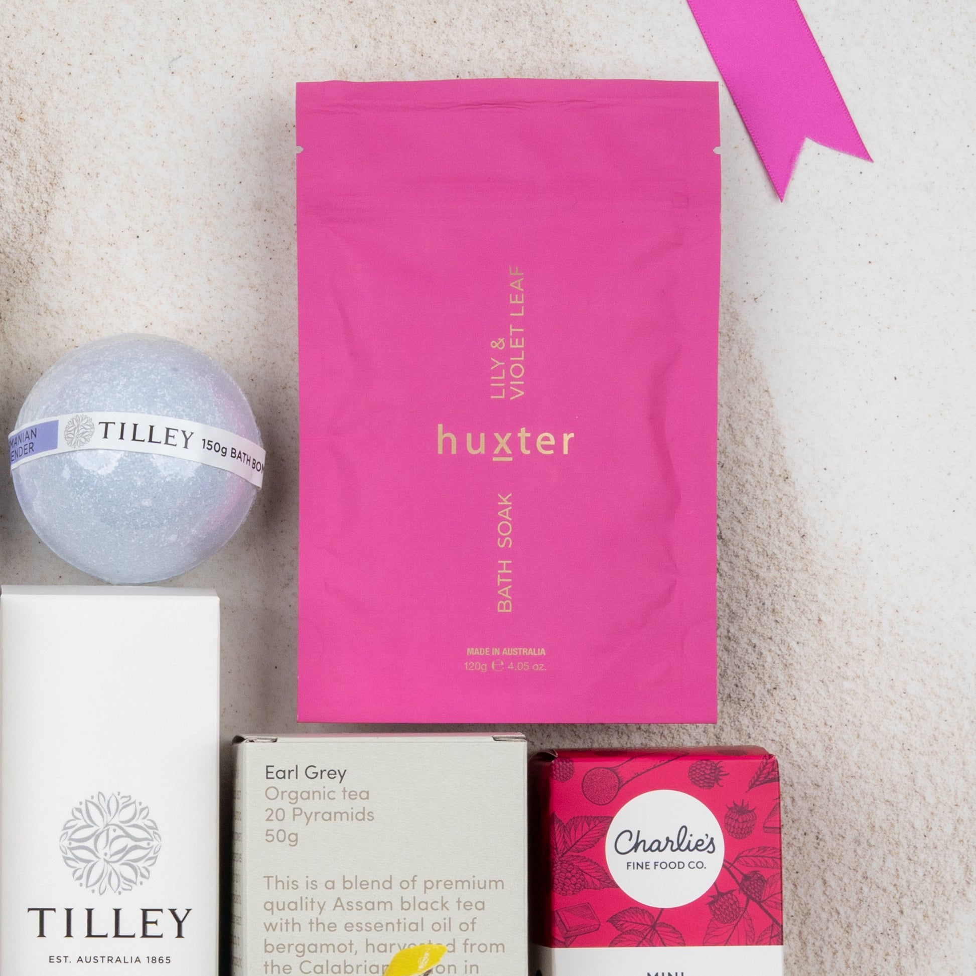 The Rejuvenation and Wellness Hamper by The Hamper Boutique Co, featuring bath soak, bath bomb, tea, and snacks, is arranged on a light surface with a pink ribbon placed in the top right corner.