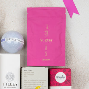 The Rejuvenation and Wellness Hamper by The Hamper Boutique Co, featuring bath soak, bath bomb, tea, and snacks, is arranged on a light surface with a pink ribbon placed in the top right corner.