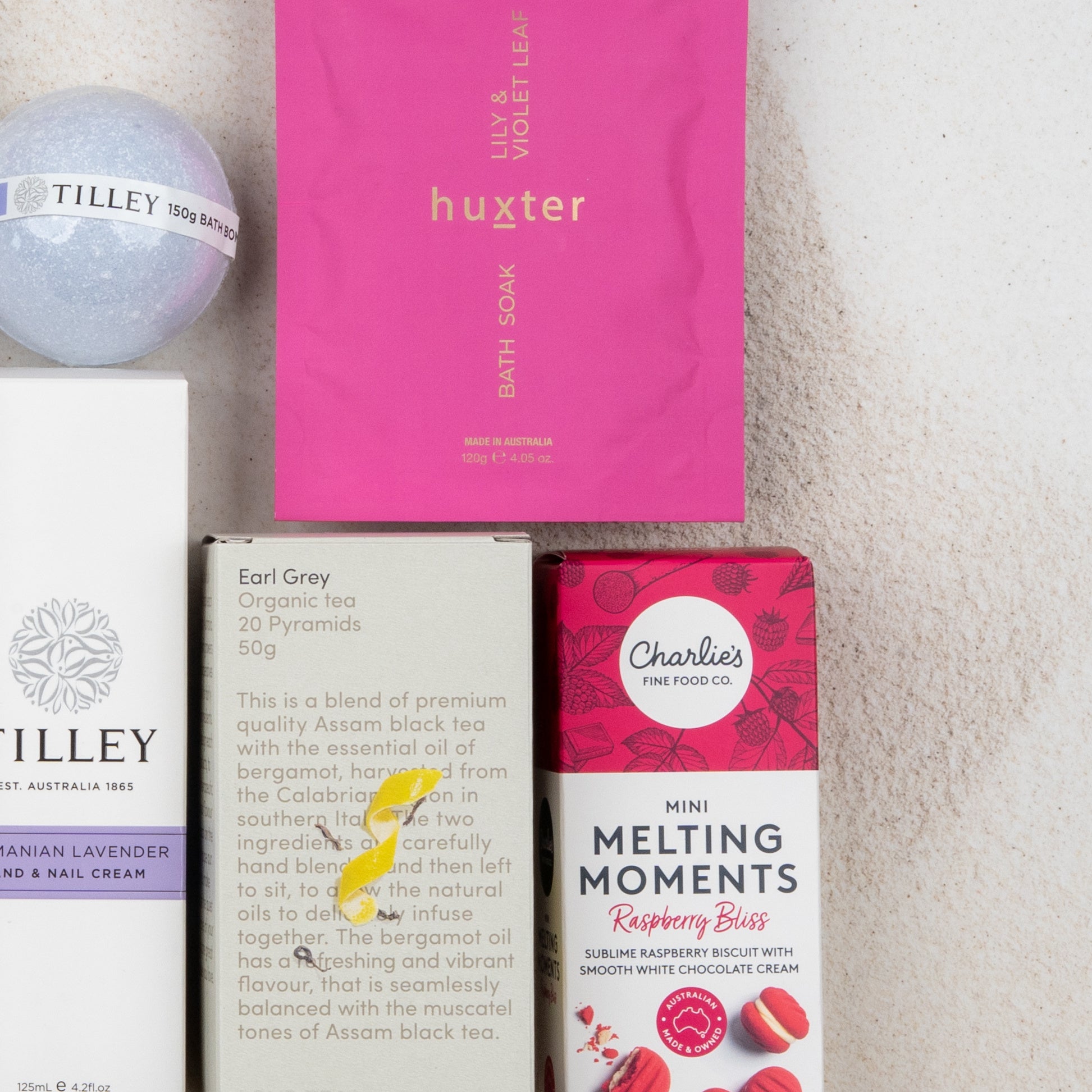 The Hamper Boutique Co's Rejuvenation and Wellness Hamper features self-care essentials, including a lavender bath bomb, pink bath soak, Earl Grey tea, and raspberry bliss melting moments cookies on a light backdrop.