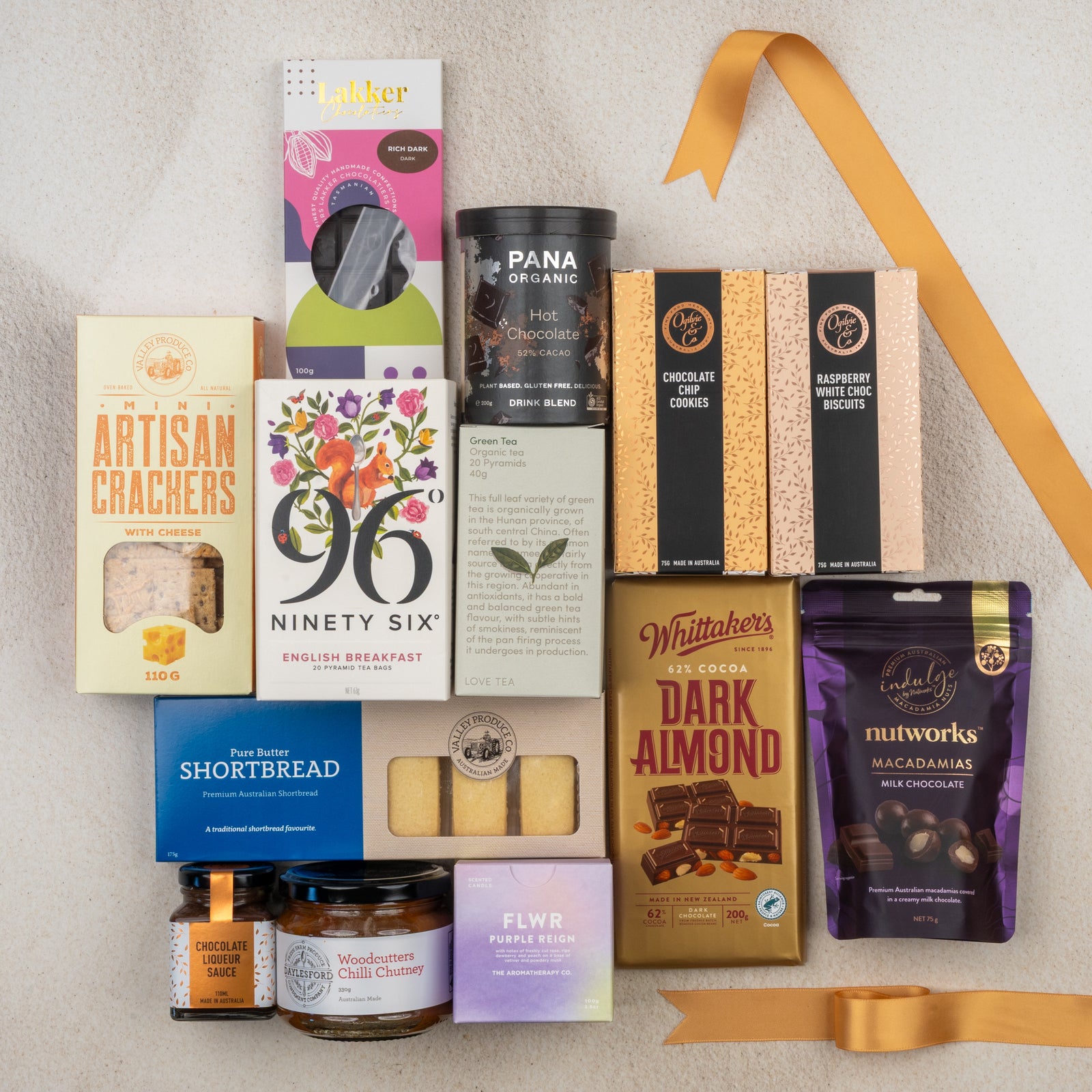 The Hamper Boutique Co’s Ultimate Tea Indulgence Hamper features gourmet snacks—crackers, biscuits, shortbread, chocolate bars, hot chocolate, chutney, and tea—elegantly arranged with a gold ribbon on a light surface.