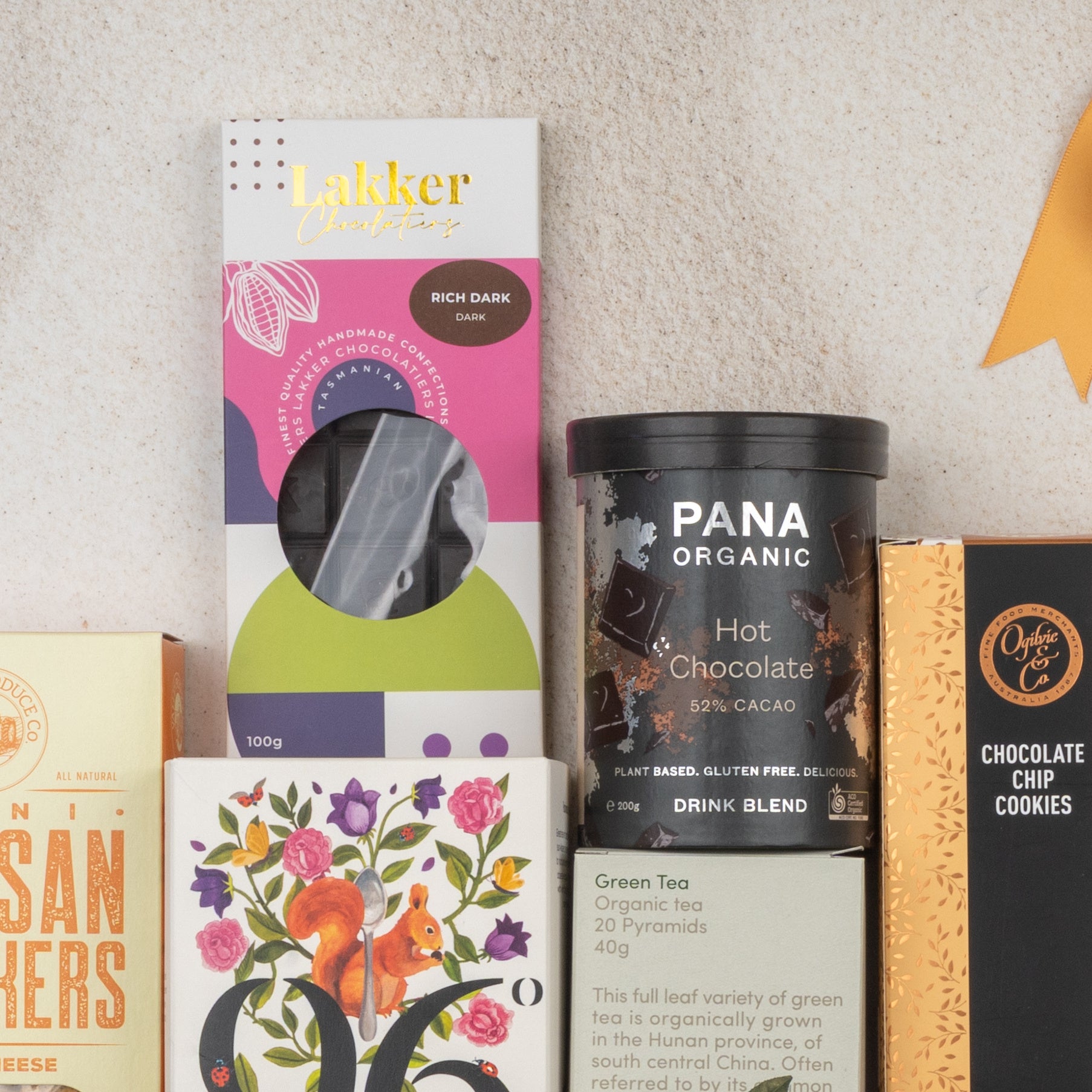 A flat lay of The Hamper Boutique Co Ultimate Tea Indulgence Hamper featuring Lakker rich dark chocolate, Pana Organic hot chocolate blend, Orange Co chocolate chip cookies, tea, and a floral bird illustrated box.