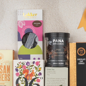 A flat lay of The Hamper Boutique Co Ultimate Tea Indulgence Hamper featuring Lakker rich dark chocolate, Pana Organic hot chocolate blend, Orange Co chocolate chip cookies, tea, and a floral bird illustrated box.