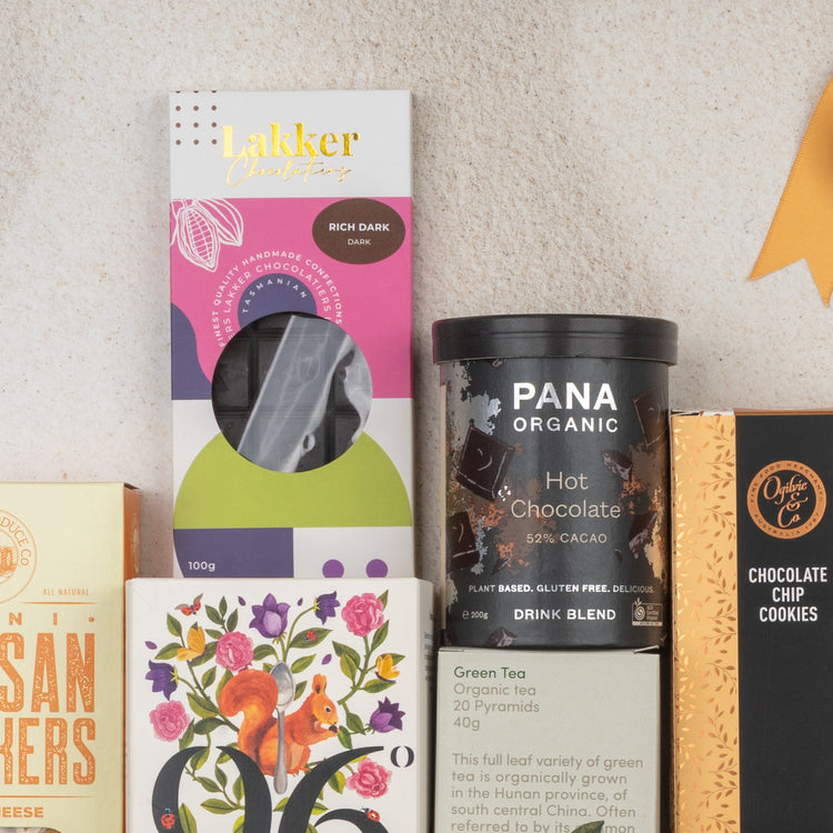A flat lay of The Hamper Boutique Co Ultimate Tea Indulgence Hamper featuring Lakker rich dark chocolate, Pana Organic hot chocolate blend, Orange Co chocolate chip cookies, tea, and a floral bird illustrated box.