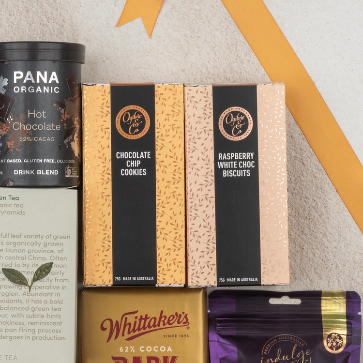 A flat lay of The Hamper Boutique Co’s Ultimate Tea Indulgence Hamper, featuring hot chocolate, chocolate chip cookies, raspberry white choc biscuits, and chocolate bars on a light surface with a gold ribbon above.