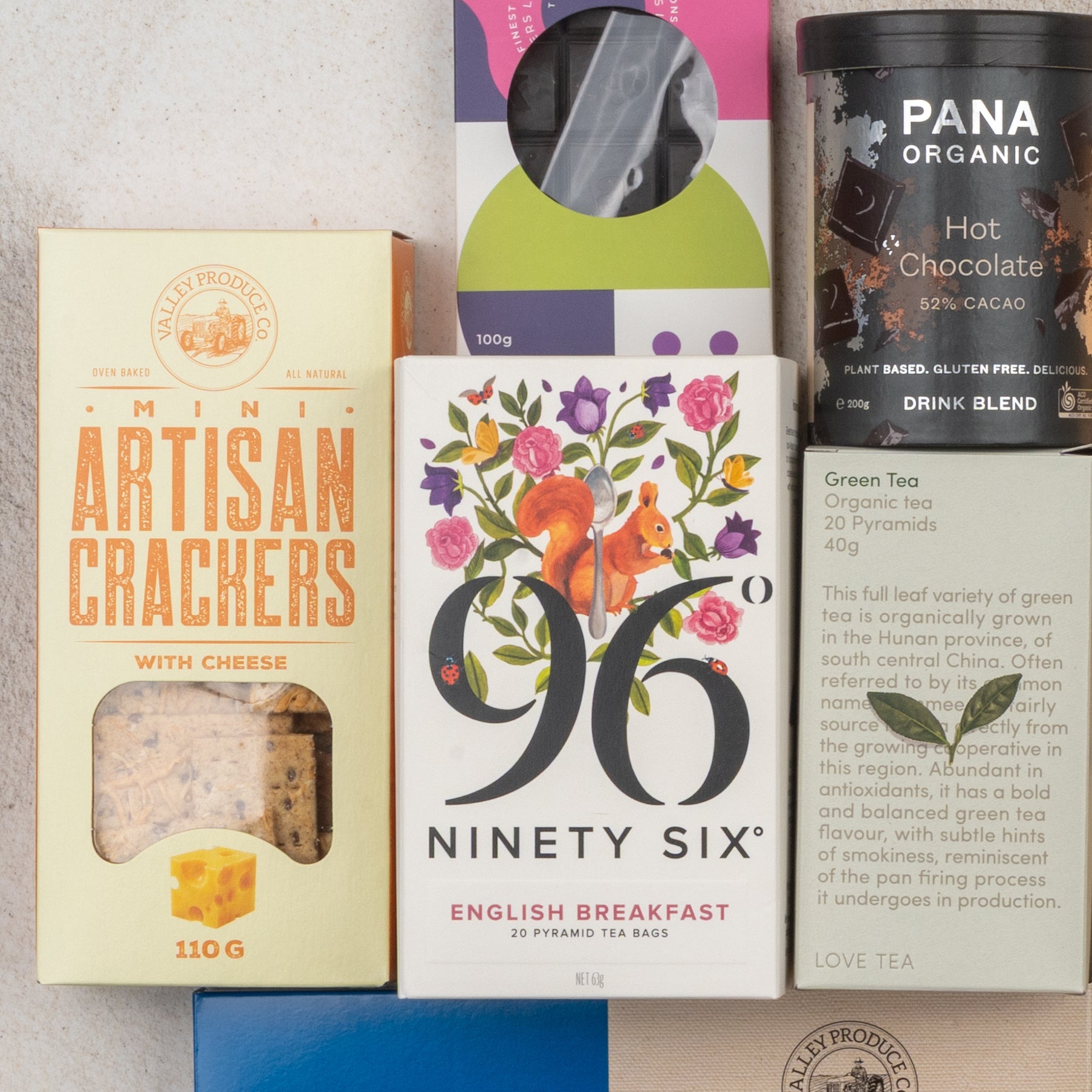 A flat lay of The Hamper Boutique Co’s Ultimate Tea Indulgence Hamper, featuring cheese mini artisan crackers, English breakfast tea with a squirrel illustration, green tea, dark chocolate, and a box with a circular window.