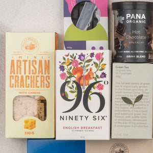 A flat lay of The Hamper Boutique Co’s Ultimate Tea Indulgence Hamper, featuring cheese mini artisan crackers, English breakfast tea with a squirrel illustration, green tea, dark chocolate, and a box with a circular window.
