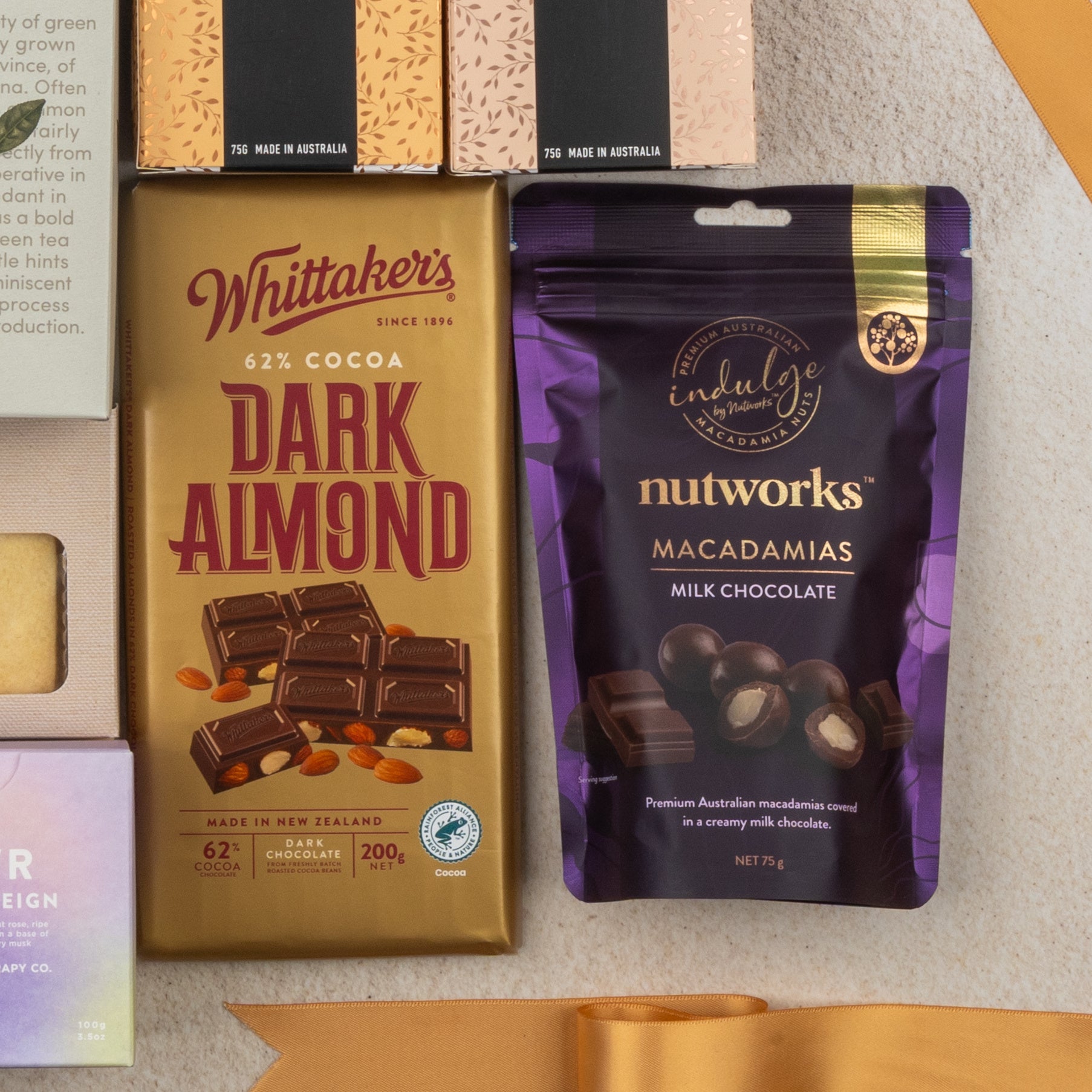 The Hamper Boutique Co's Ultimate Tea Indulgence Hamper is displayed on a beige surface, accompanied by other packaged treats and a gold ribbon.