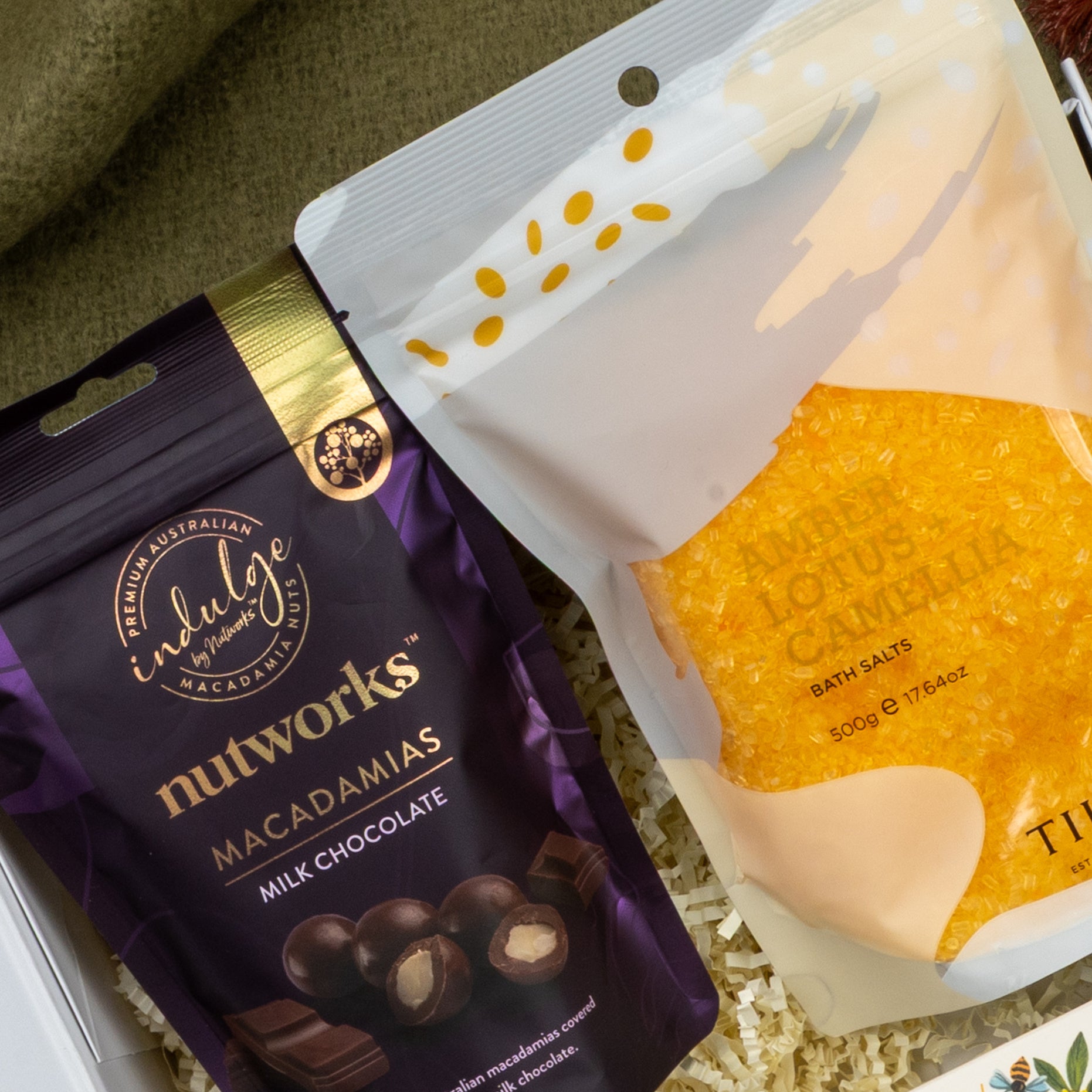 A Wellness Home Entertaining Hamper by The Hamper Boutique Co displays Nutworks milk chocolate macadamias in a purple bag beside Timsbee lotus camellia bath salts in yellow-orange packaging on shredded paper.