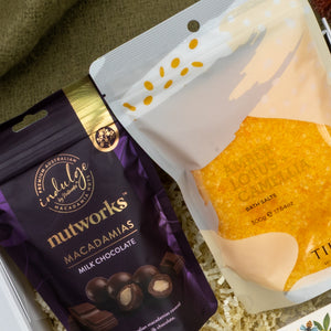 A Wellness Home Entertaining Hamper by The Hamper Boutique Co displays Nutworks milk chocolate macadamias in a purple bag beside Timsbee lotus camellia bath salts in yellow-orange packaging on shredded paper.