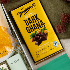 A Wellness Home Entertaining Hamper from The Hamper Boutique Co sits on a tabletop near a green leaf, boxed yellow item, mint-green packaged item, and a 72% cocoa Whittaker’s Dark Ghana chocolate bar described as a special blend.