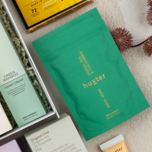 A flat lay of The Hamper Boutique Co's Wellness Home Entertaining Hamper, featuring spa products like green tea & cucumber bath soak, bamboo hand cream, a chocolate bar, and assorted items on a beige surface.