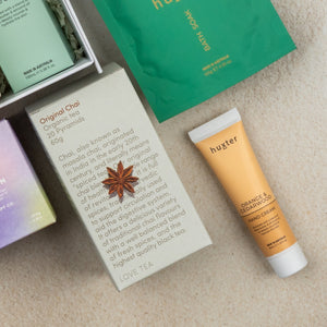 Flat lay of The Hamper Boutique Co Wellness Home Entertaining Hamper, featuring a box of Original Chai tea with an anise star, Huxter hand cream, and green and purple packaged items on a light textured surface.