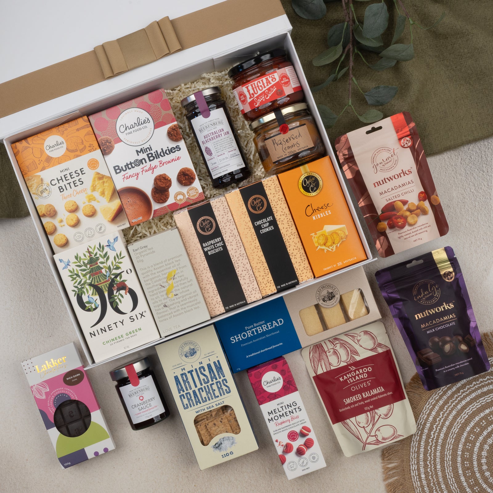 The Hamper Boutique Co's Tea Time Gourmet Hamper features an elegant selection of crackers, cheese bites, nuts, cookies, chocolates, jam, and preserves in a gift box presented on a white background with green foliage accents.