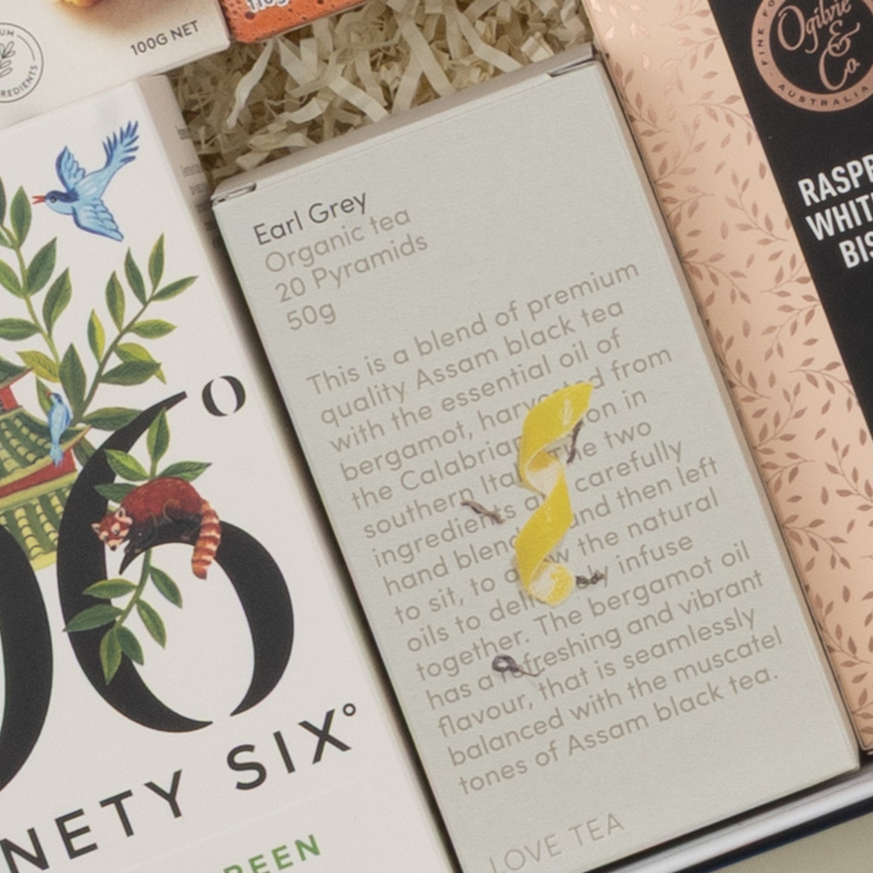 A close-up of The Hamper Boutique Co’s Tea Time Gourmet Hamper shows a gray Earl Grey Organic Tea box with blend details and a yellow bird illustration. Other boxes in the hamper display decorative patterns.