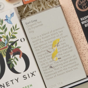 A close-up of The Hamper Boutique Co’s Tea Time Gourmet Hamper shows a gray Earl Grey Organic Tea box with blend details and a yellow bird illustration. Other boxes in the hamper display decorative patterns.