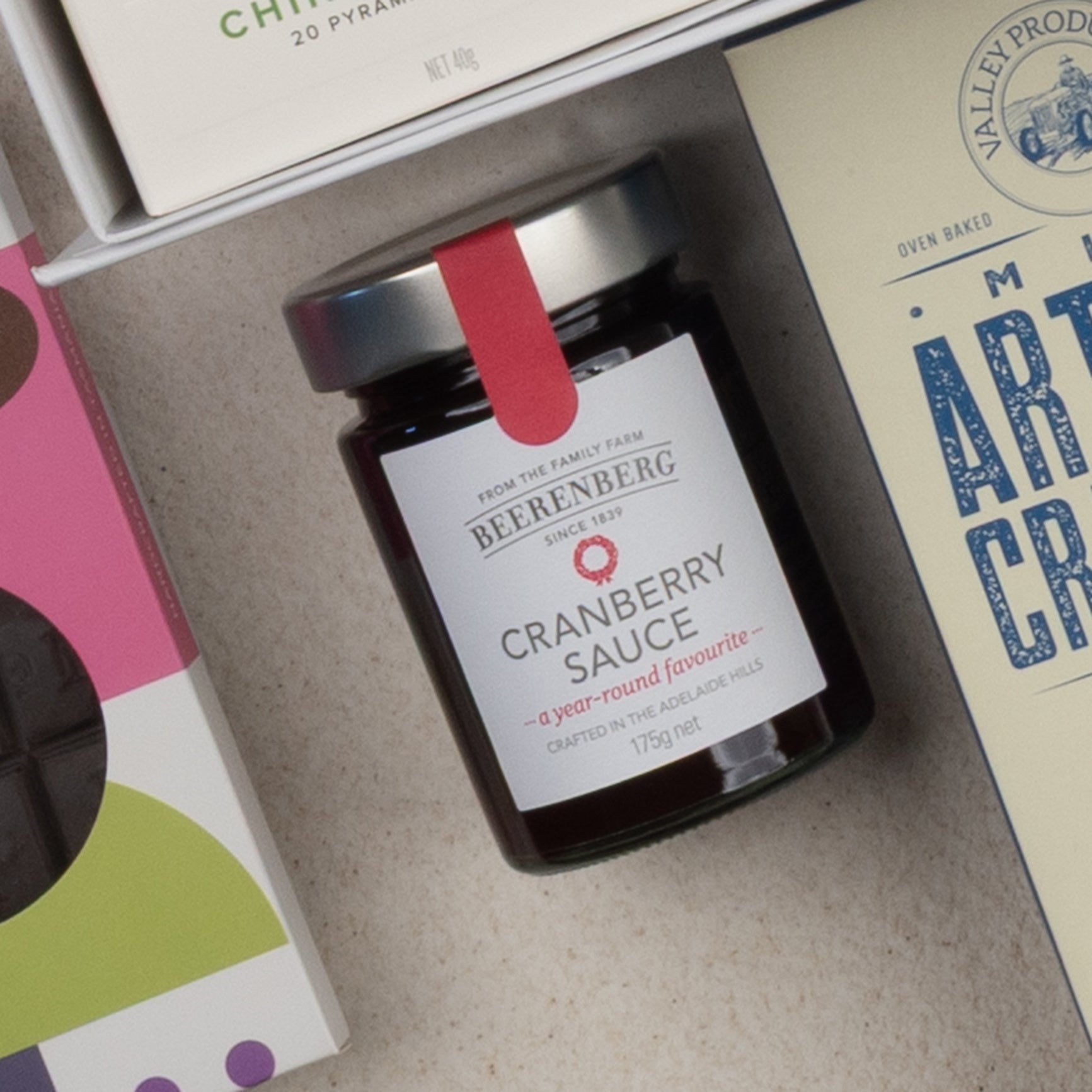 The Tea Time Gourmet Hamper by The Hamper Boutique Co is displayed from above, featuring a selection of packaged food items such as a box of milk artisan crackers and a purple polka-dotted box.