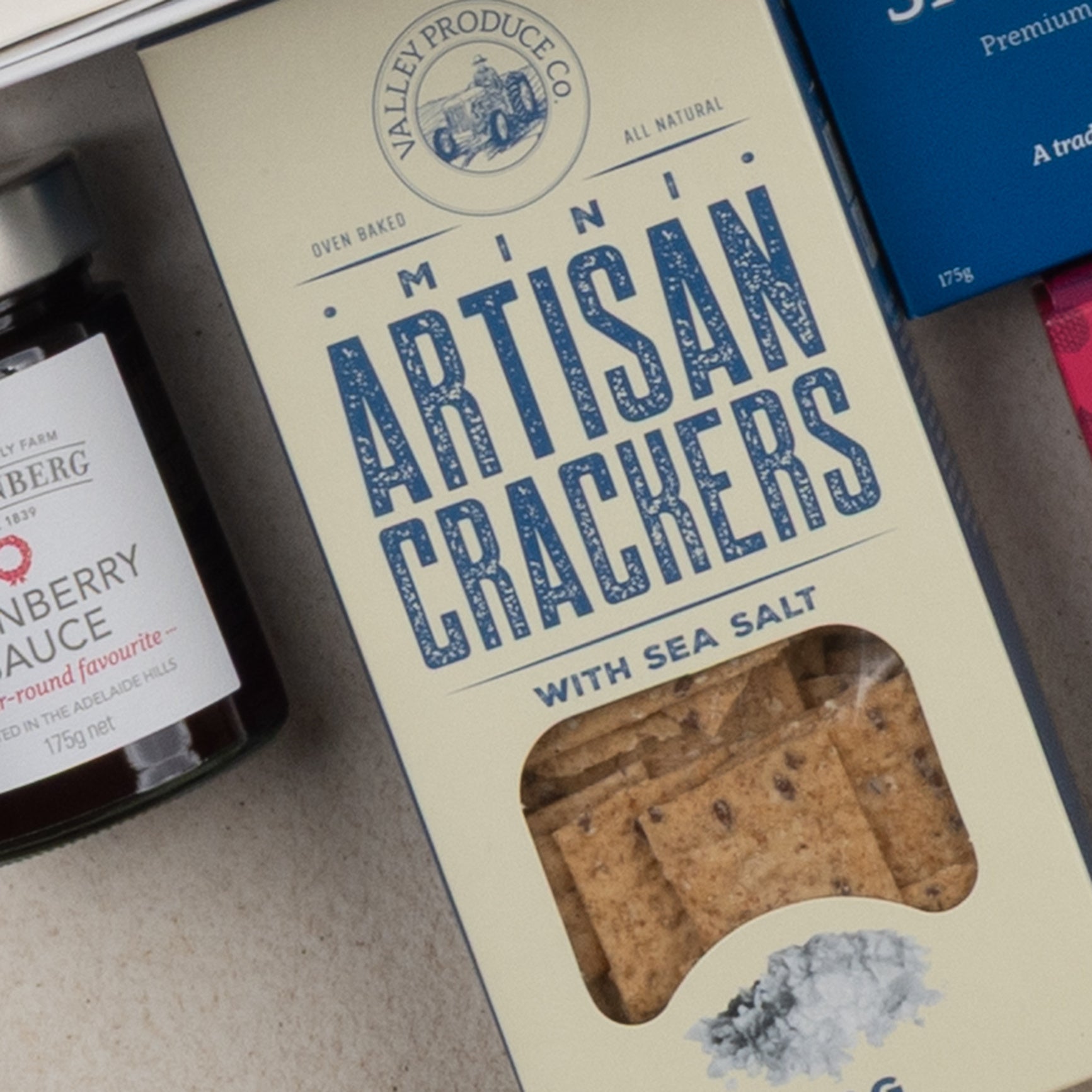 A close-up of The Hamper Boutique Co's Tea Time Gourmet Hamper, showing a box with crackers visible through a window, placed beside a jar of cranberry preserve and part of another blue package.