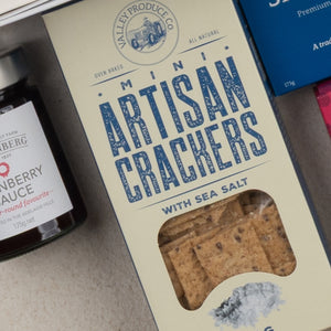 A close-up of The Hamper Boutique Co's Tea Time Gourmet Hamper, showing a box with crackers visible through a window, placed beside a jar of cranberry preserve and part of another blue package.