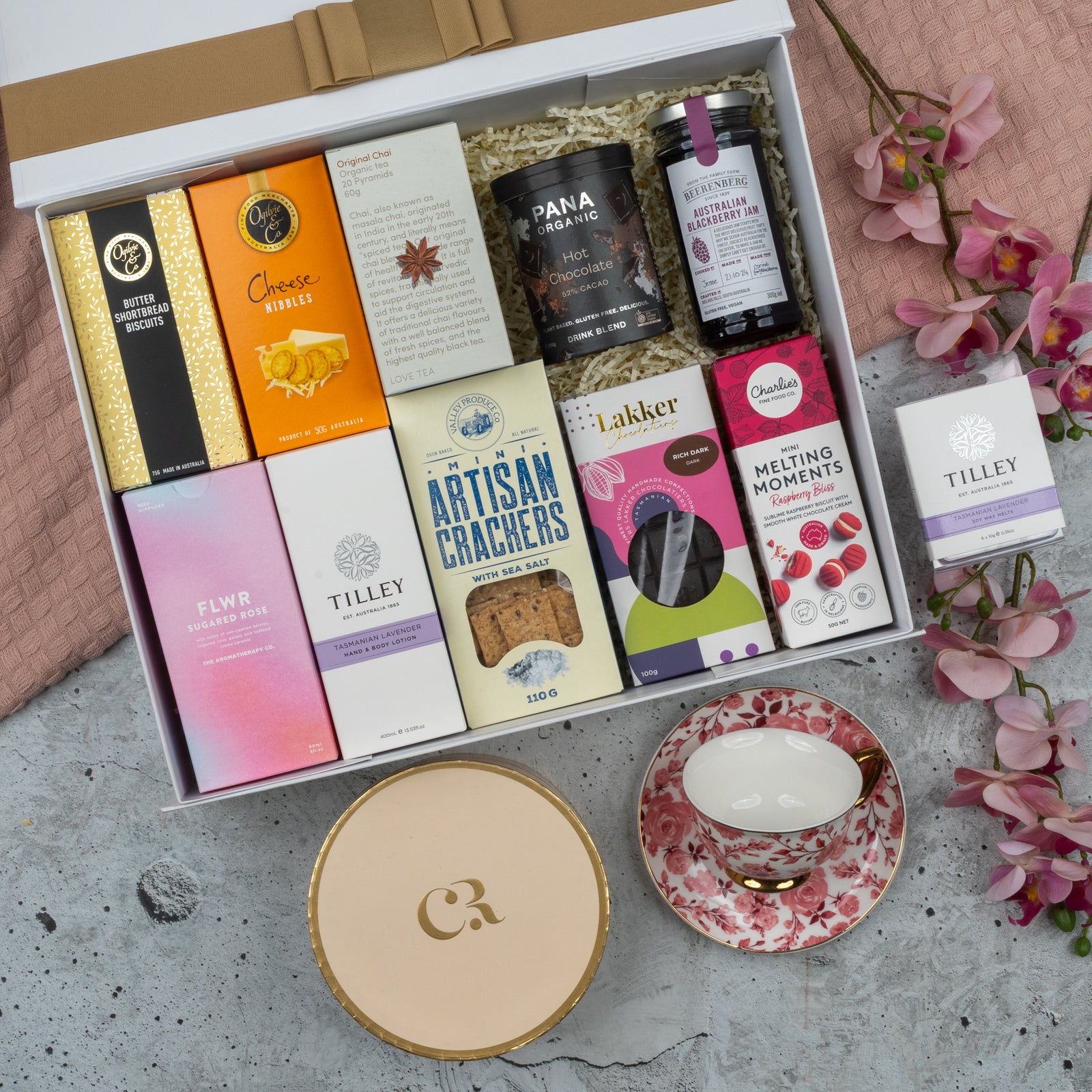 The Hamper Boutique Co's Premium Tea Experience Hamper features gourmet snacks, tea, chocolates, crackers, and a pink floral teacup with saucer, beautifully arranged with pink flowers and a gold-ribboned lid.