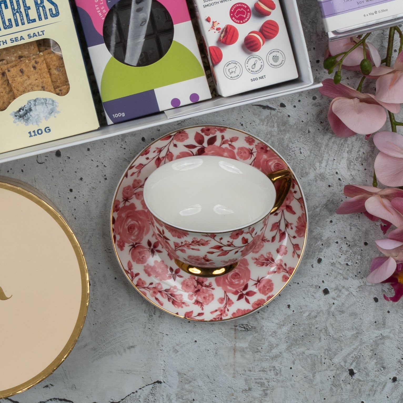 The Hamper Boutique Co’s Premium Tea Experience Hamper features a pink and white floral teacup with gold accents, displayed on a gray surface and surrounded by snack boxes and pink flowers.