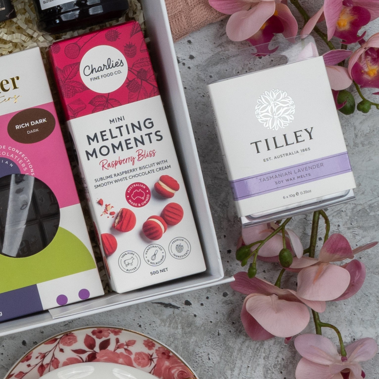The Hamper Boutique Co Premium Tea Experience Hamper, featuring chocolates, raspberry bliss cookies, and Tasmanian lavender soy wax melts, styled with pink orchids on a gray surface.