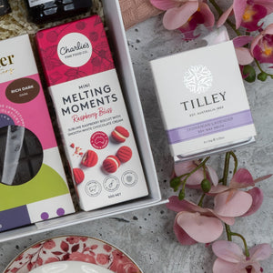 The Hamper Boutique Co Premium Tea Experience Hamper, featuring chocolates, raspberry bliss cookies, and Tasmanian lavender soy wax melts, styled with pink orchids on a gray surface.