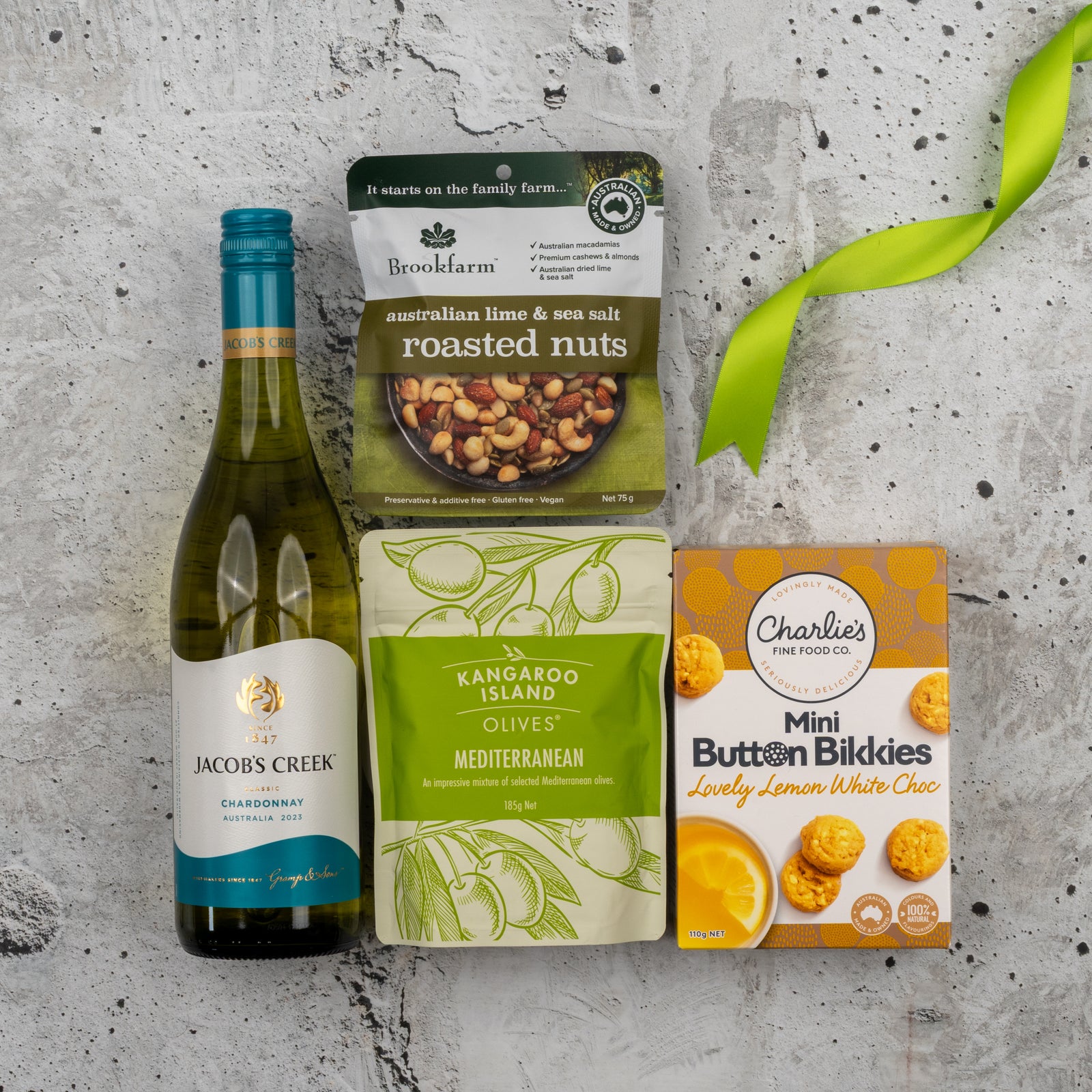 The Hamper Boutique Co's Gourmet Treats Gift Hamper features Chardonnay, roasted nuts, Mediterranean olives, and mini white chocolate cookies arranged on a gray stone surface with a green ribbon in the corner.