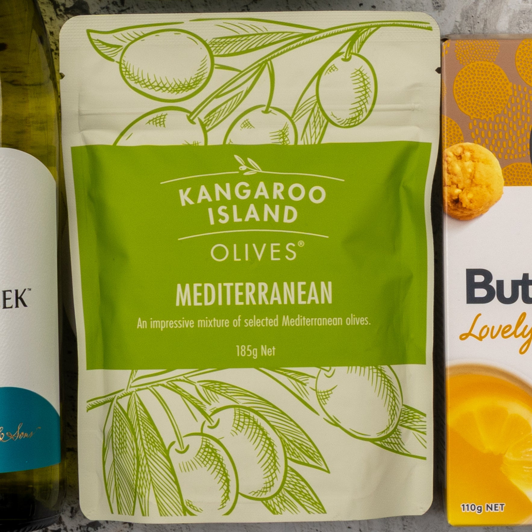 A green and cream pouch from The Hamper Boutique Co Gourmet Treats Gift Hamper sits between a bottle and box, featuring illustrations of olives and olive branches on its packaging.
