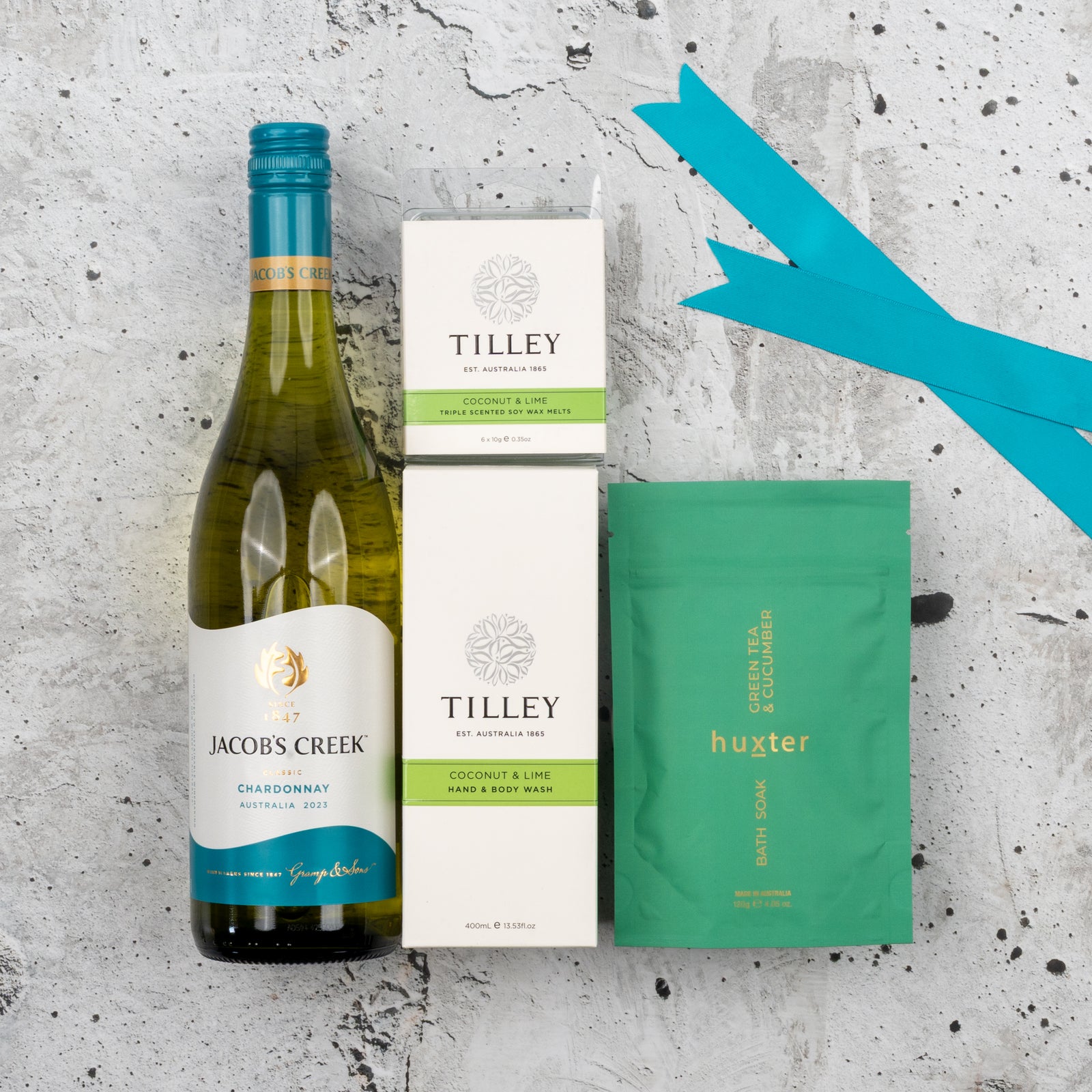 The Hamper Boutique Co's Spa Day Retreat Gift Hamper is displayed on a textured gray surface with teal ribbons, featuring Jacob’s Creek Chardonnay, Tilley coconut & lime bath products, and a green Huxter bath soak packet.