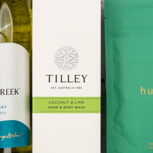 Close-up of The Hamper Boutique Co’s Spa Day Retreat Gift Hamper, featuring a bottle of white wine, Tilley coconut & lime hand and body wash, and a partially visible green package with orange text.