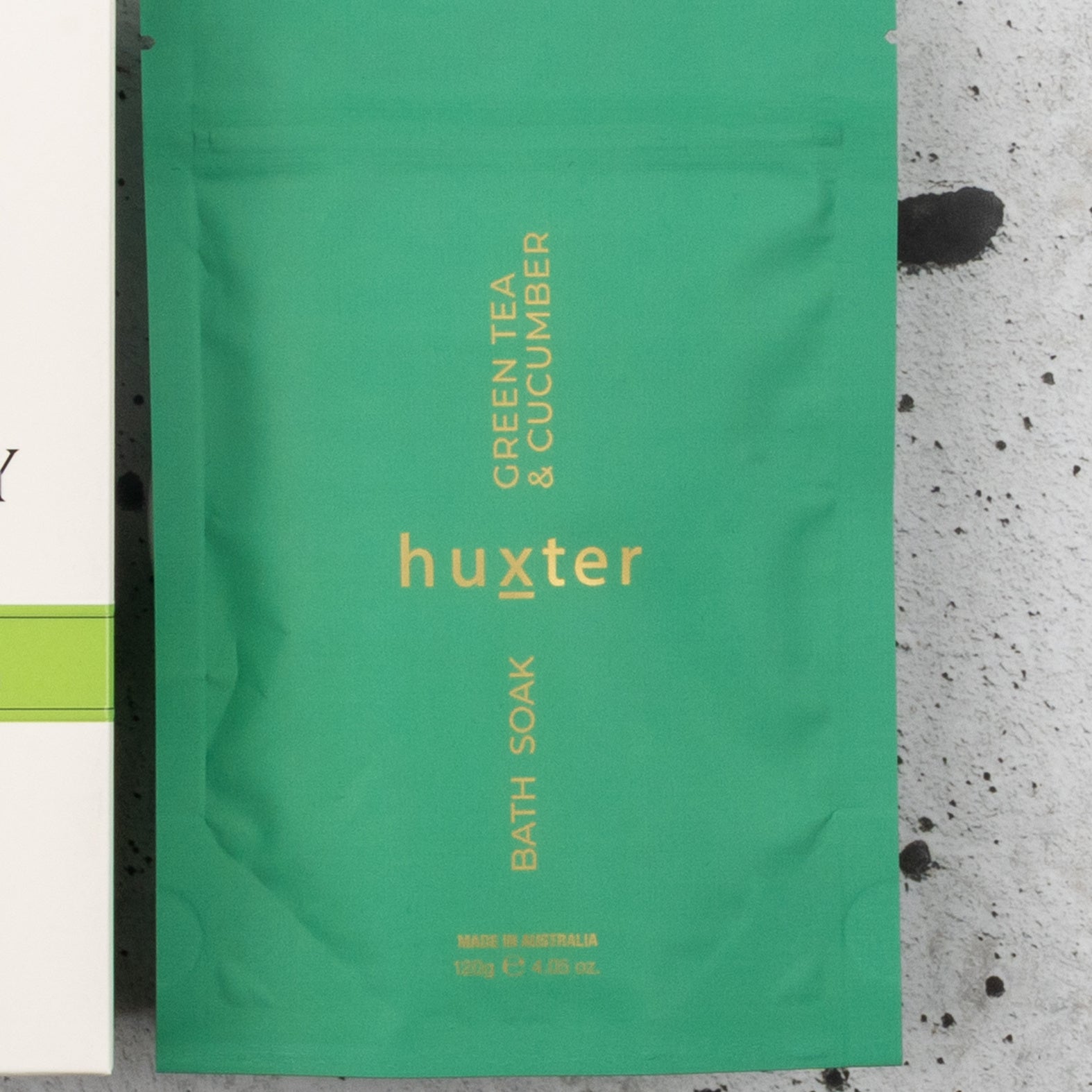 A packet from The Hamper Boutique Co's Spa Day Retreat Gift Hamper, with yellow "huxter" text and "Green Tea & Cucumber Bath Soak," displayed on a light gray, dark-speckled surface.