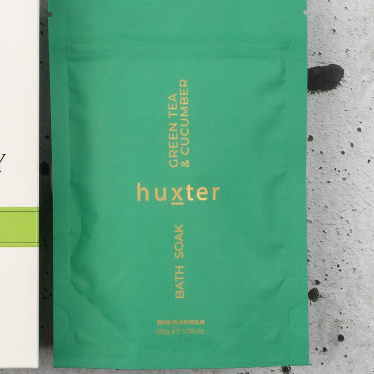 A packet from The Hamper Boutique Co's Spa Day Retreat Gift Hamper, with yellow "huxter" text and "Green Tea & Cucumber Bath Soak," displayed on a light gray, dark-speckled surface.