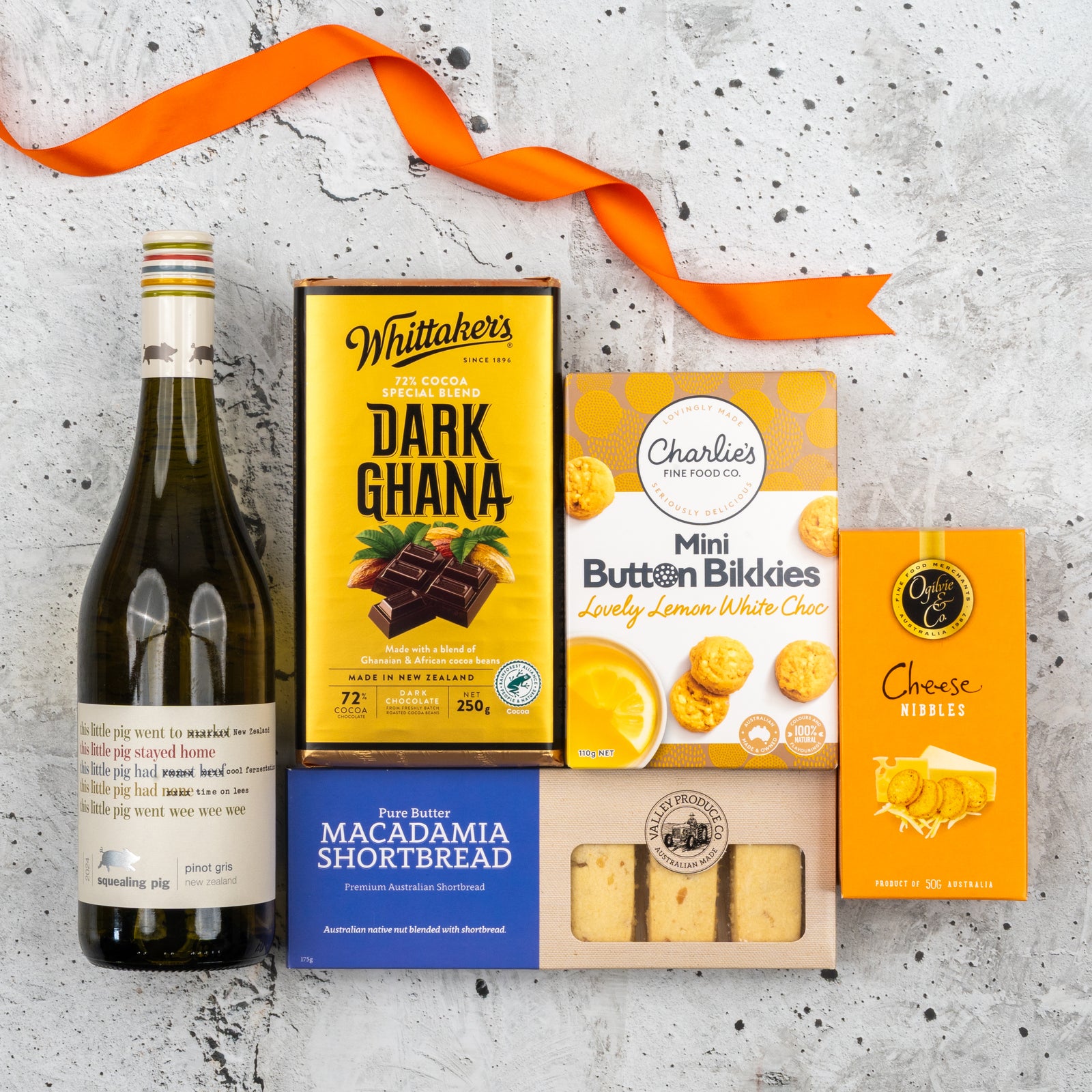The Hamper Boutique Co’s Classic Elegance Gift Hamper features white wine, dark chocolate, butter macadamia shortbread, mini white choc button cookies, cheese nibbles, and an orange ribbon on a light gray textured surface.