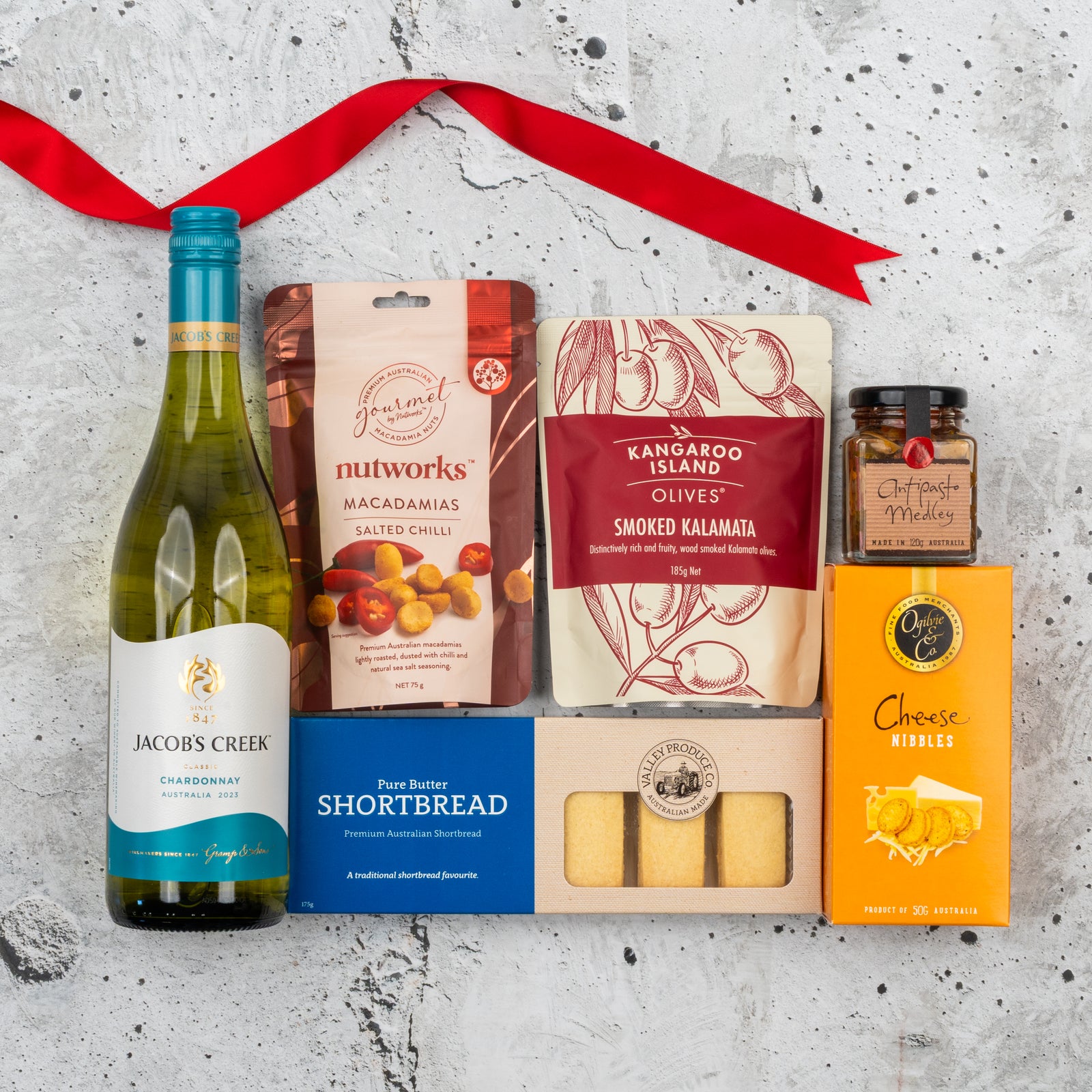 The Hamper Boutique Co Gourmet Getaway Gift Hamper on a gray surface with a red ribbon, featuring Jacob’s Creek Chardonnay, salted chili macadamias, smoked kalamata olives, oat & seed cheese nibbles, butter shortbread, and chutney.